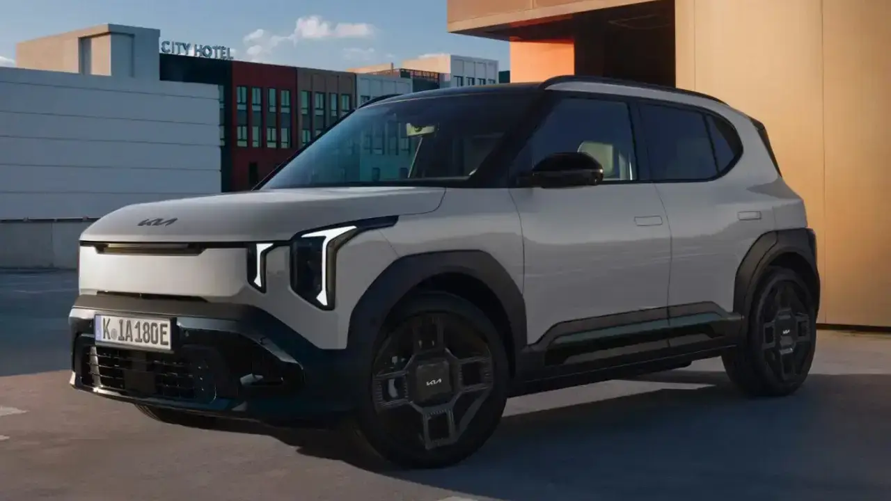 Der neue vollelektrische Kia EV3 in Hellgrau auf einem urbanen Parkdeck. Das kompakte Elektro-SUV zeigt seine markante Front mit T-förmigen LED-Scheinwerfern vor einer Stadtkulisse
