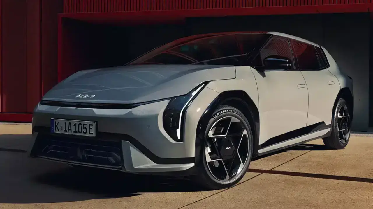 Schräge Frontansicht des grauen Kia EV4 Hatchback Elektroautos. Das kompakte E-Modell der GT-Line zeigt seine markanten vertikalen LED-Scheinwerfer und futuristischen Alufelgen vor einer roten Gebäudefassade