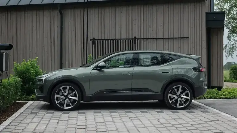 Seitenansicht des Polestar 3 800V SE Elektro-SUVs in Salbeigrün beim Ladevorgang an einer Wallbox. Die Profil-Ansicht zeigt die coupéhafte Linienführung und sportliche 7-Speichen-Felgen vor einer modernen Holzfassade