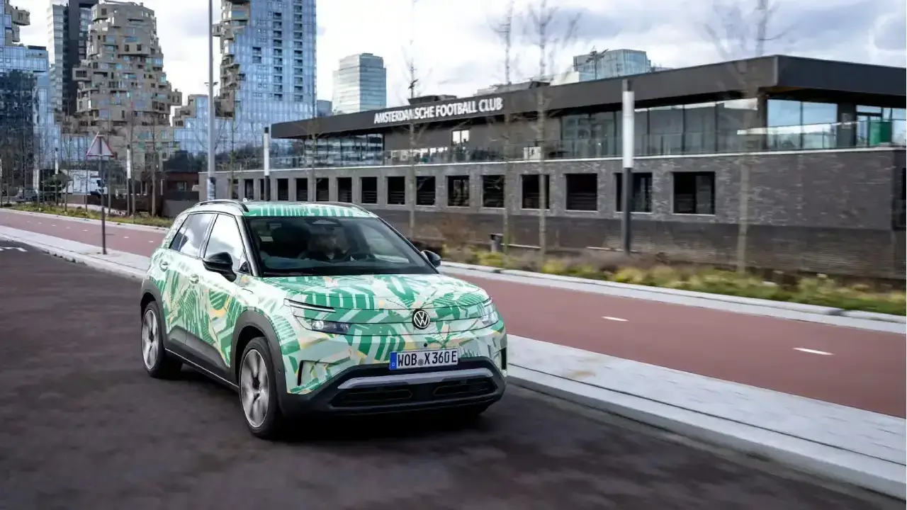 Schräge Frontansicht eines getarnten VW ID. Cross Elektro-SUV-Prototyps auf der Straße. Der kompakte Erlkönig zeigt eine auffällige grüne Tarnfolie im Palmenblatt-Design vor einer modernen Stadtkulisse
